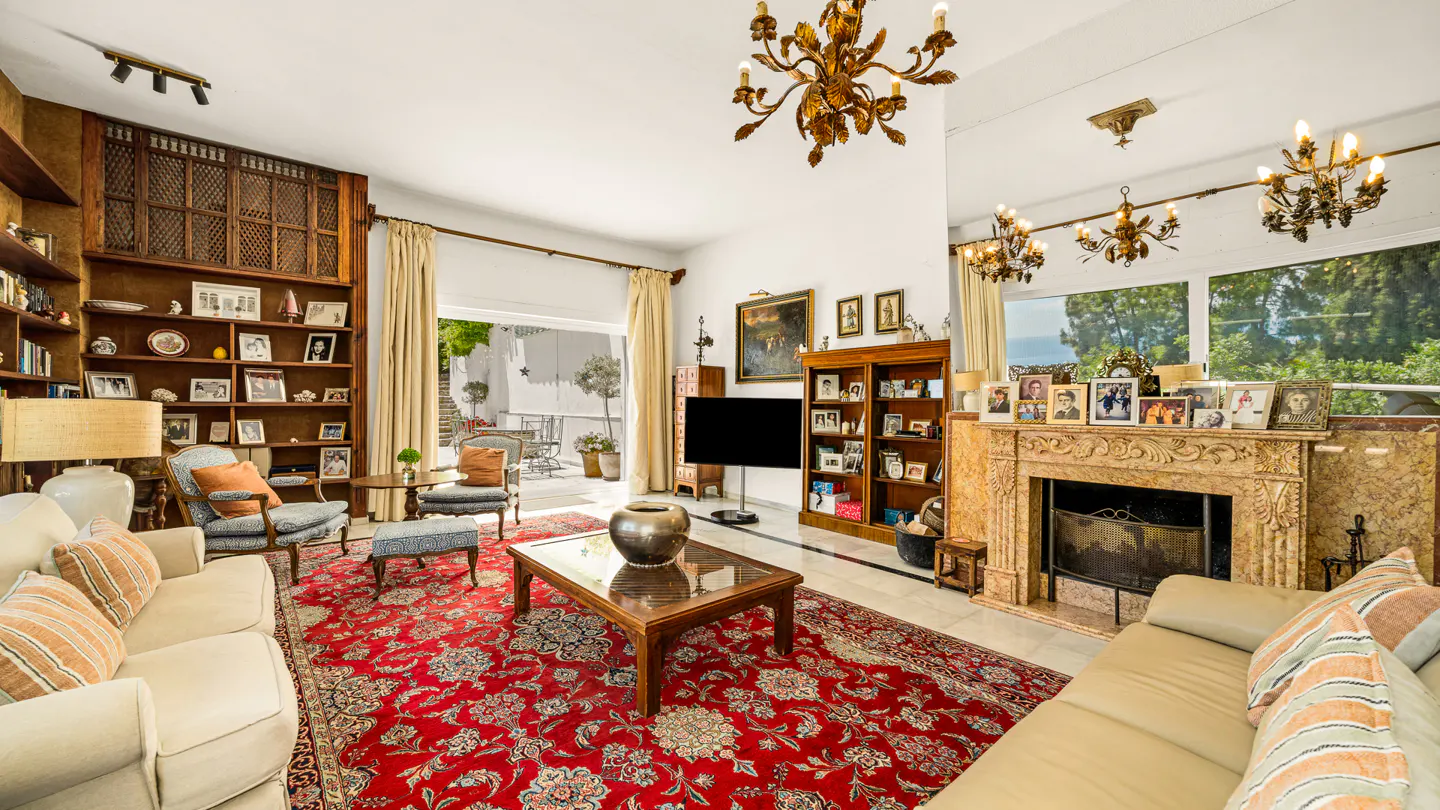 Bright living room with sofas, a red floral rug, and a fireplace. Bookshelves are filled with photos and decor. Chandeliers hang from the ceiling.