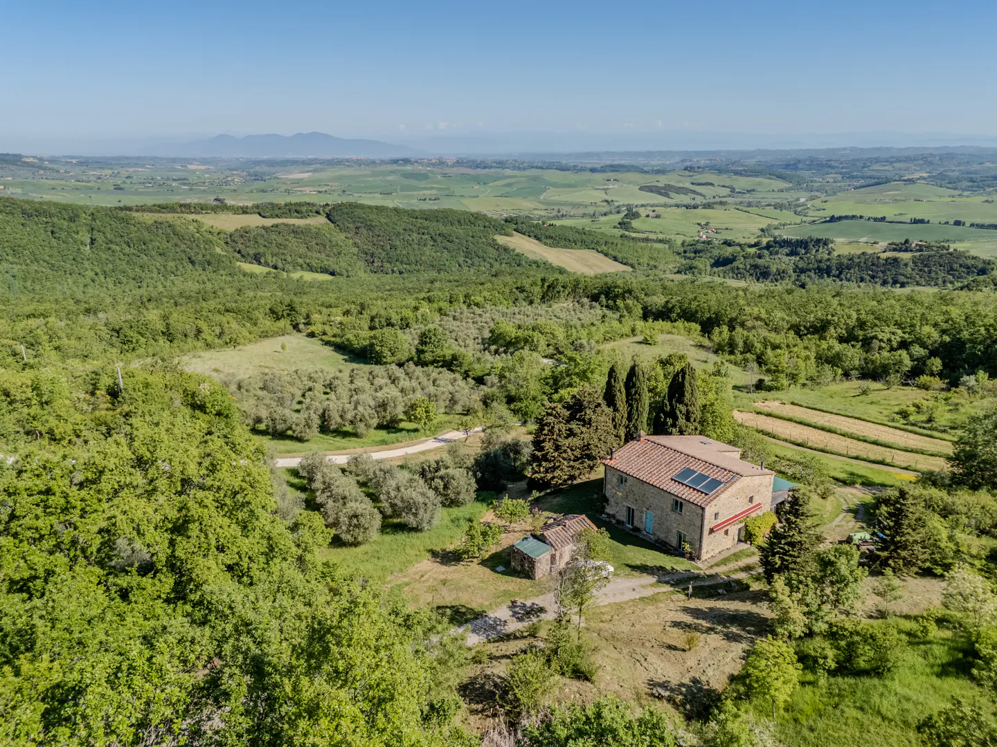 Aerial view of a stone house with a red tile roof and solar panels, surrounded by green trees and rolling hills.