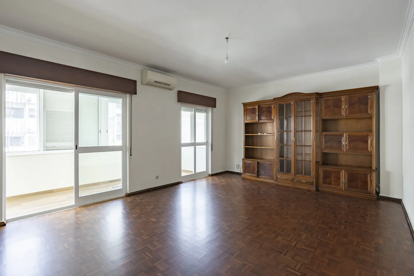 Bright, empty room with parquet floors, white walls, and a large wooden bookcase. Sliding glass doors lead to a balcony.