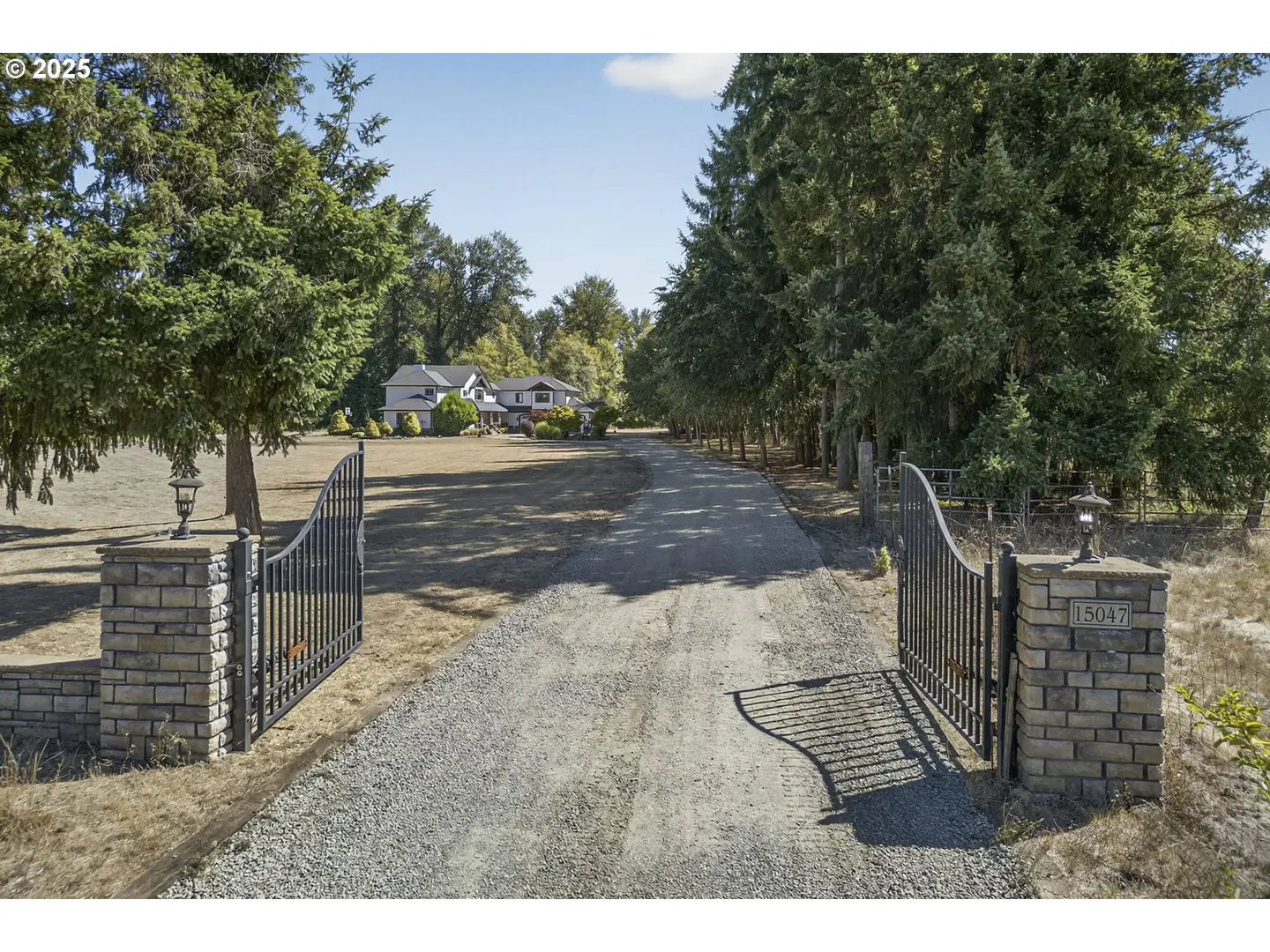 Gravel driveway leads to a white house, framed by trees and an open black metal gate with stone pillars.