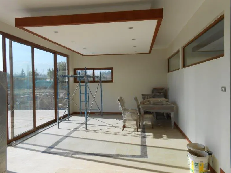 Bright, empty room with large windows, a blue scaffold, and furniture covered in plastic wrap. The ceiling has a brown wood trim.