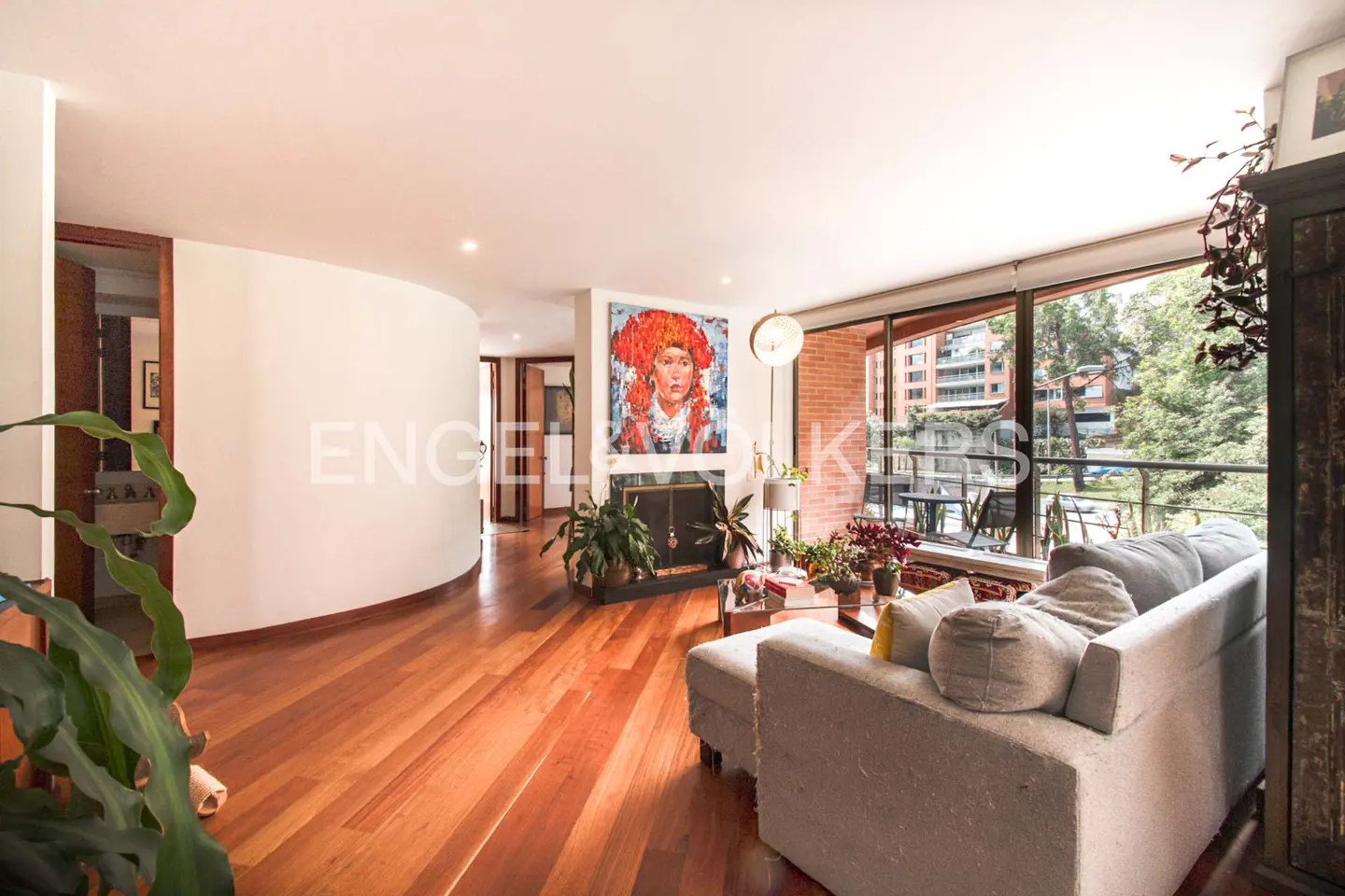 Bright living room with hardwood floors, a gray sofa, a fireplace with a large portrait, and floor-to-ceiling windows.
