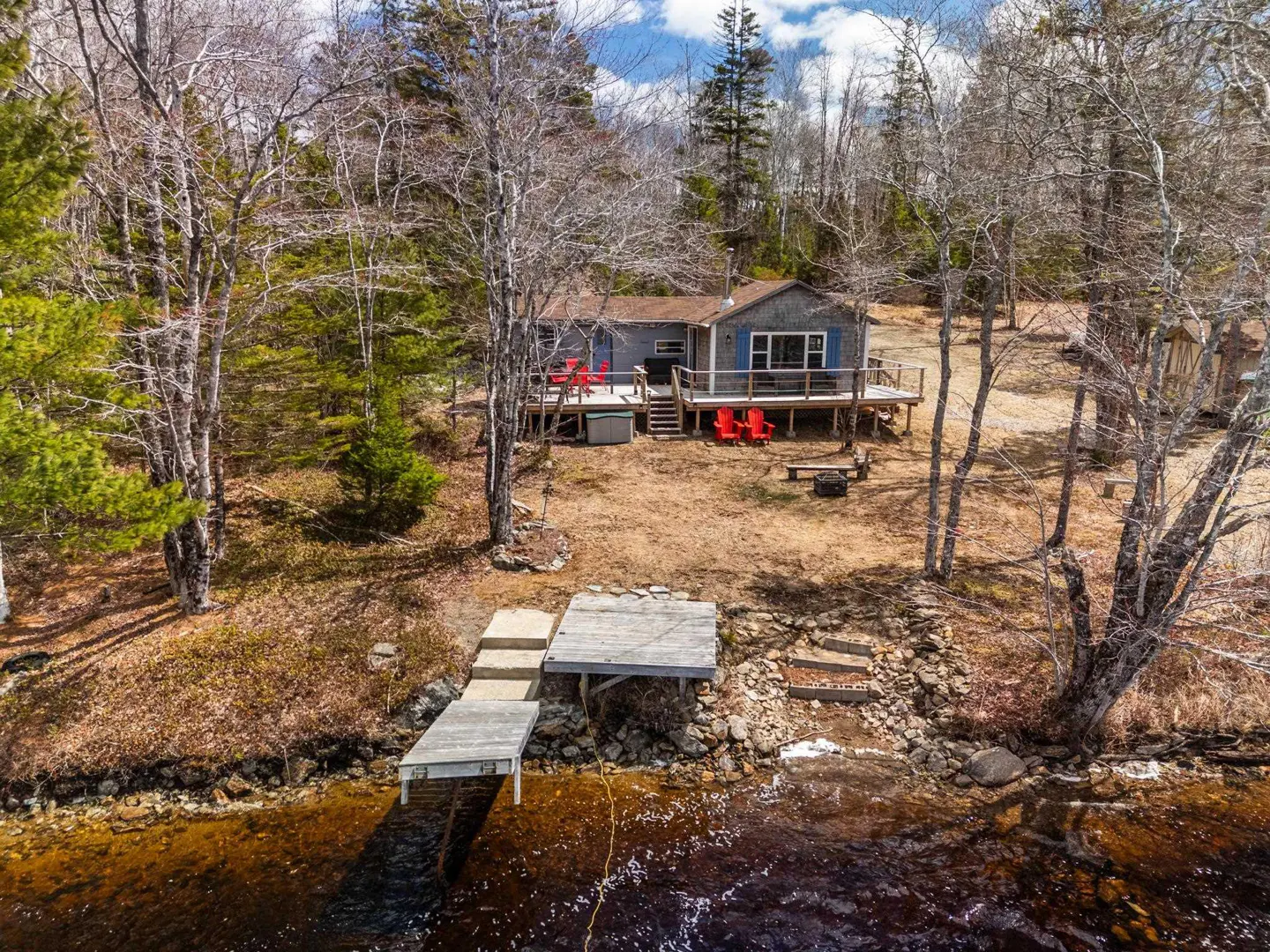 Aerial view of a gray lake house with a wooden deck, red chairs, and a dock leading to the water. Trees surround the house.