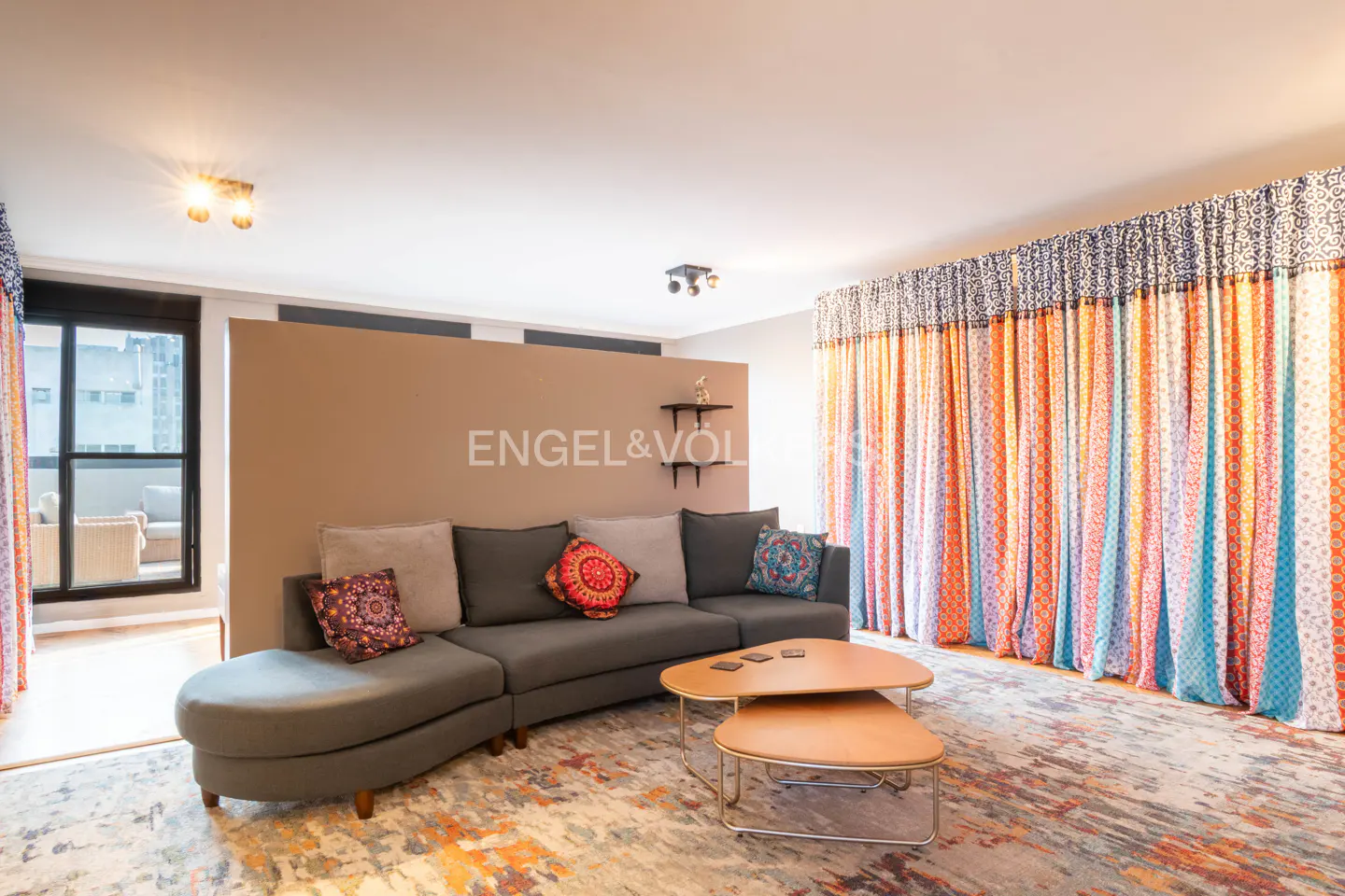 Living room with gray sectional sofa, patterned pillows, and two-tiered coffee table on a colorful rug. Bright curtains and balcony view.