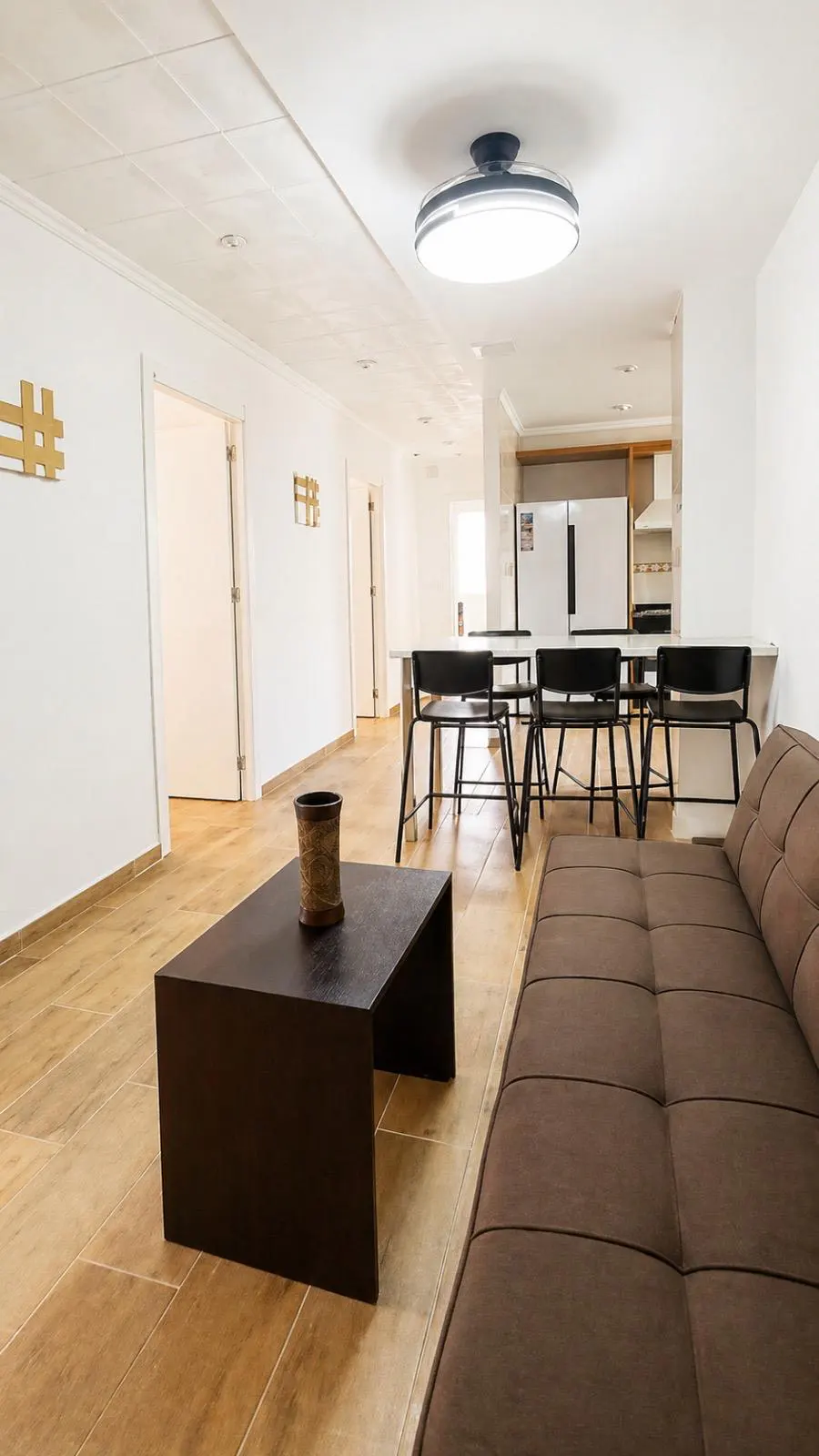 Bright, open-concept living space with a brown sofa, dark coffee table, and a kitchen in the background. White walls and light wood floors.