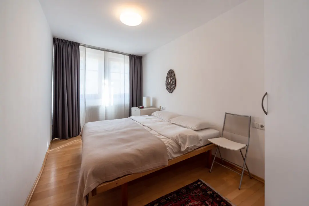 Bright bedroom with a wooden bed frame, white linens, and a beige blanket. A white chair sits on a red rug. Gray curtains frame a window.