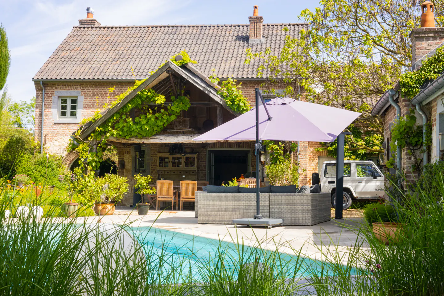 Exterior view of a brick house with a pool, patio furniture, and a purple umbrella. Green plants and a white car are also visible.