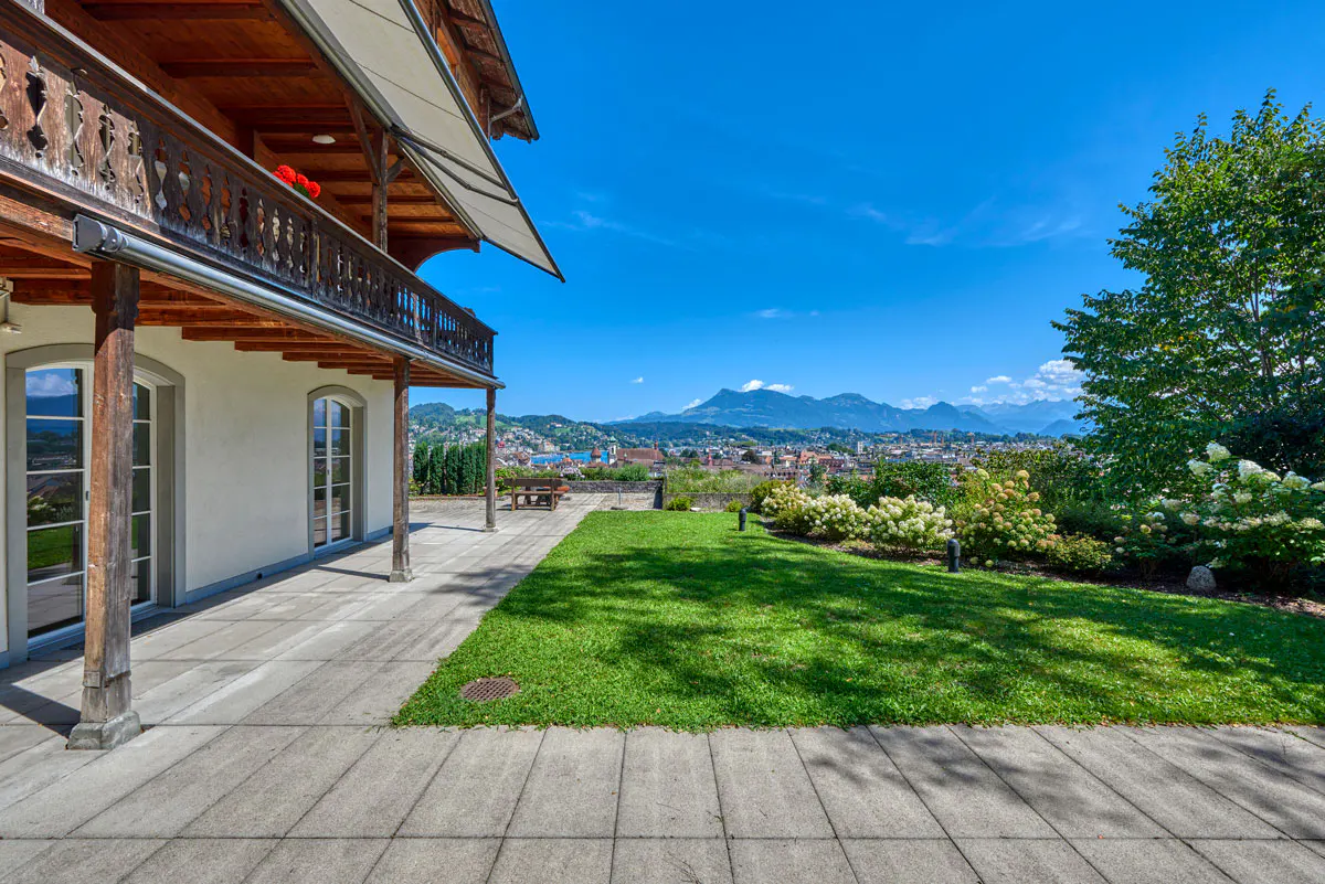 Exterior view of a house with a balcony, green lawn, and a city view with mountains in the background under a blue sky.