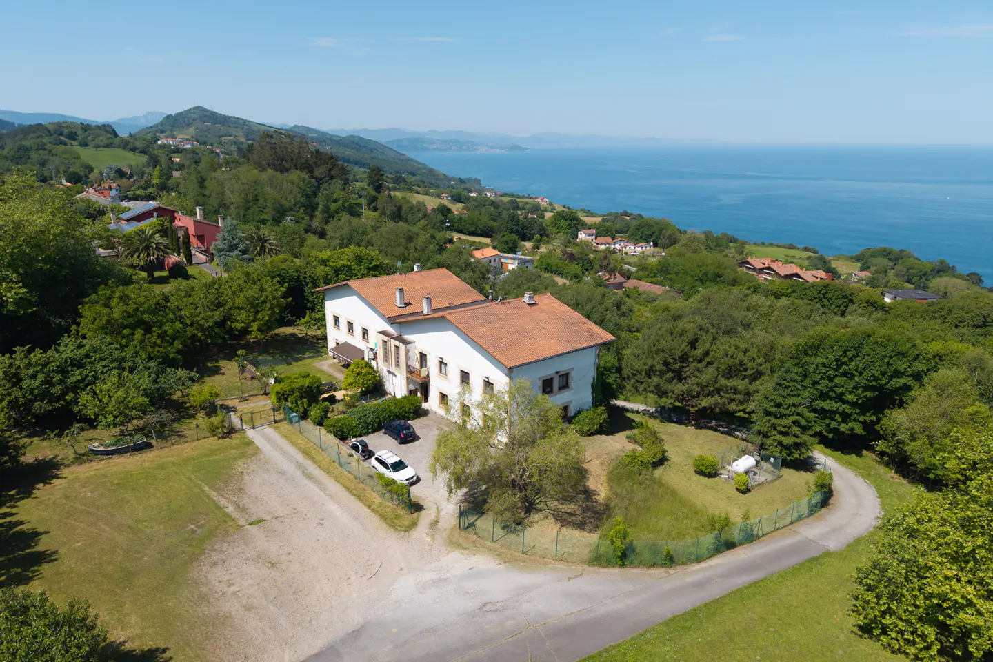 Aerial view of a white two-story house with a red tile roof, surrounded by green trees and a blue ocean in the background.