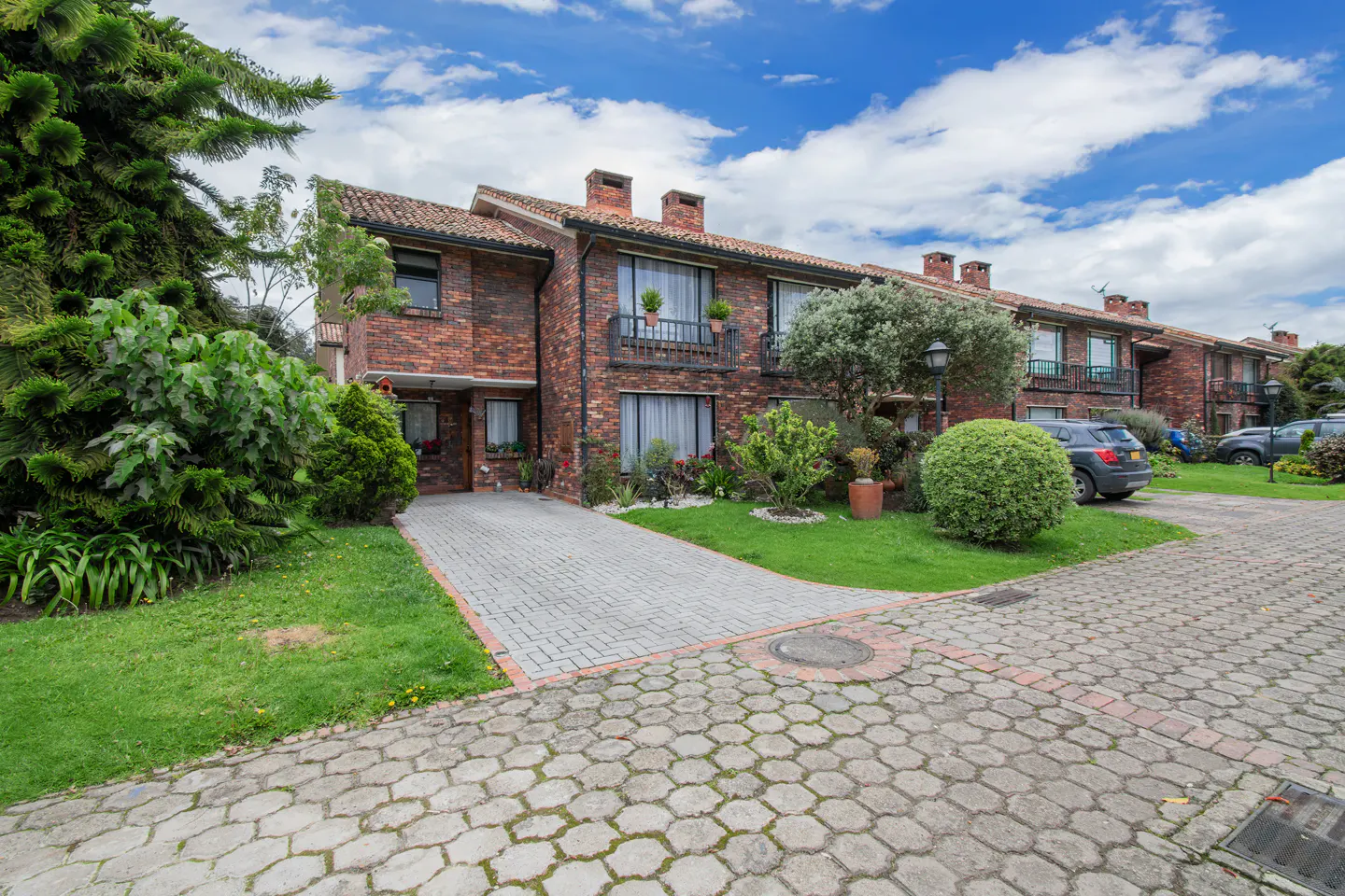 Brick townhouses with red tile roofs line a paved driveway under a blue, cloudy sky. Green lawns and bushes surround the homes.