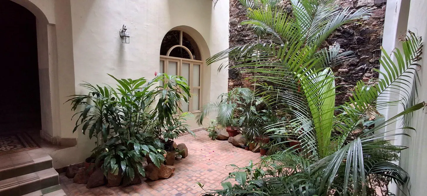 Indoor courtyard with brick floor, stone wall, and lush green plants. Arched window and wall sconce add architectural detail.