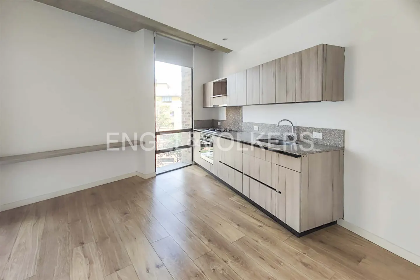 A bright, modern kitchen with light wood cabinets, granite countertops, and wood floors. A large window offers a view of greenery.