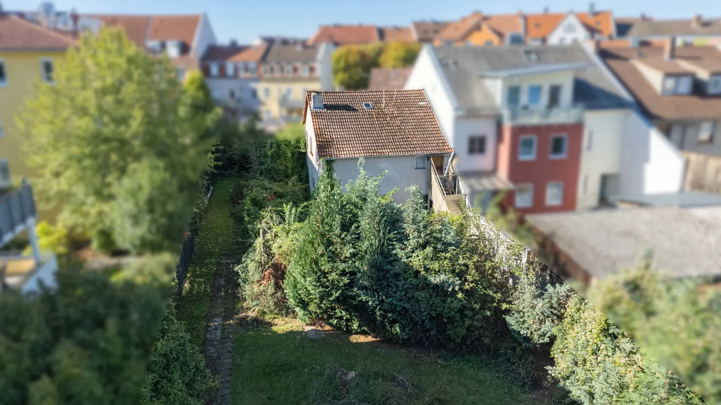 Aerial view of a small, light-colored house with a red-tiled roof, surrounded by lush green trees and a long, narrow garden. Other houses are blurred in the background.