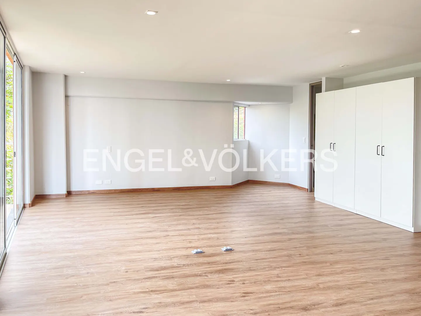 An empty room with light wood floors, white walls, and a large white wardrobe. A window and glass doors let in natural light. Engel & Völkers logo on the wall.