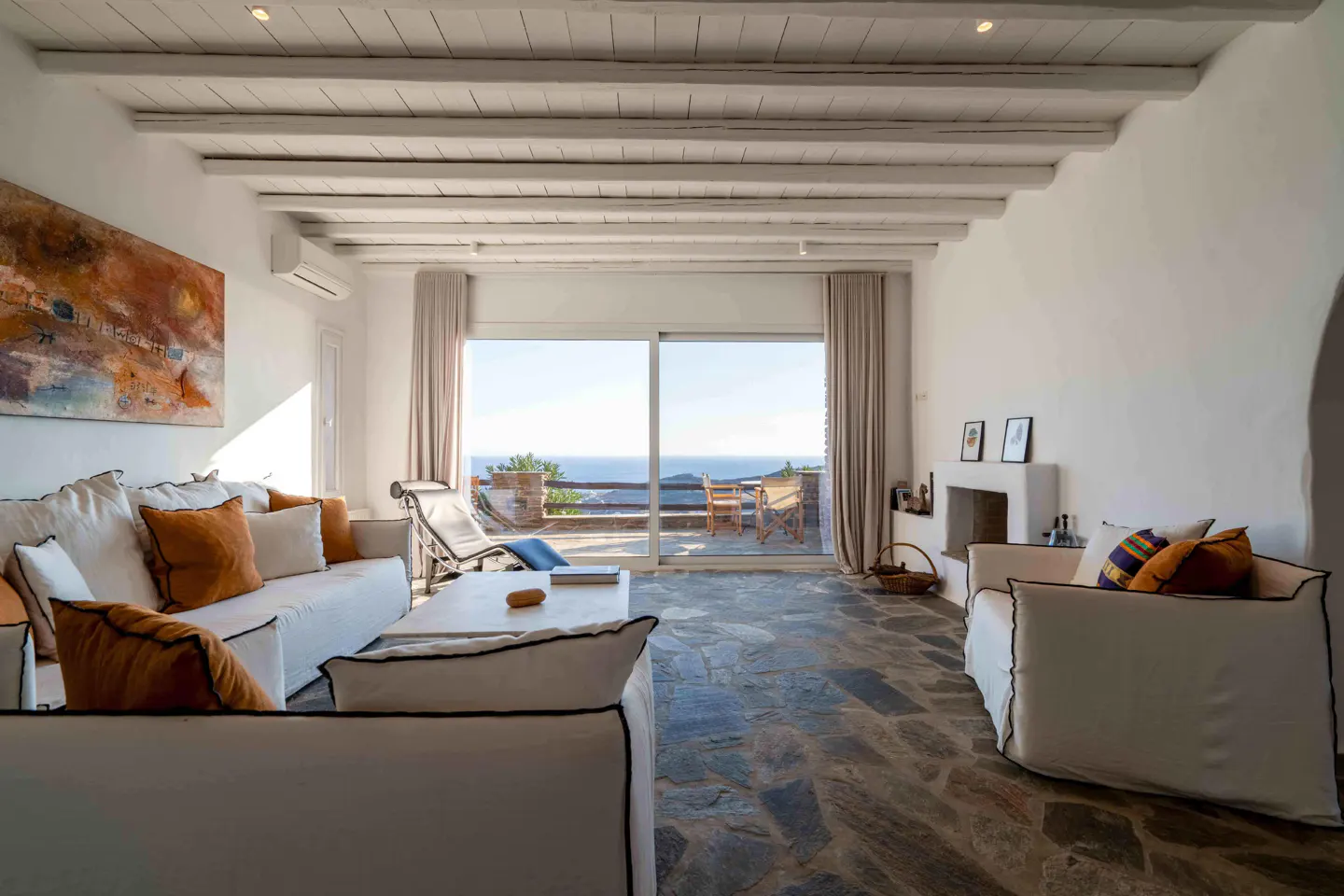 Living room with white sofas, stone floor, and white beamed ceiling, featuring a large window overlooking the ocean.