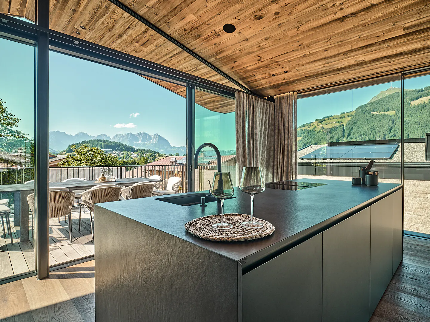Modern kitchen with wood ceiling, black island, and mountain view. Table and chairs visible on the deck through sliding glass doors.