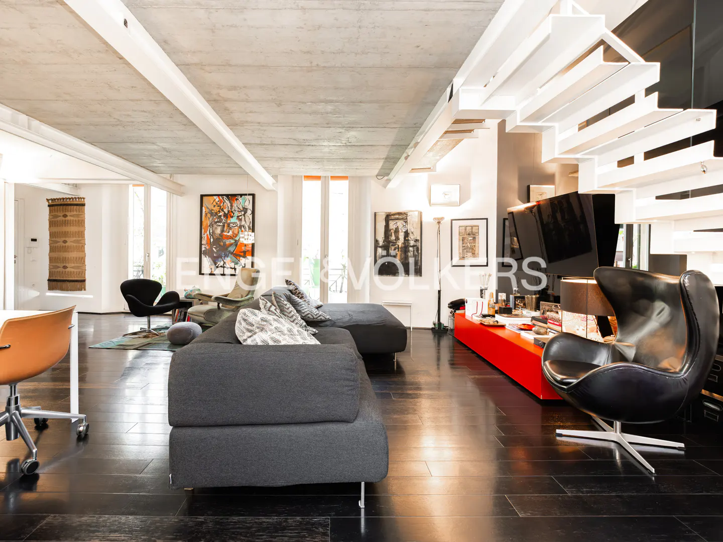 Modern living room with gray sofa, black wood floors, and white staircase. Art and furniture add pops of color.