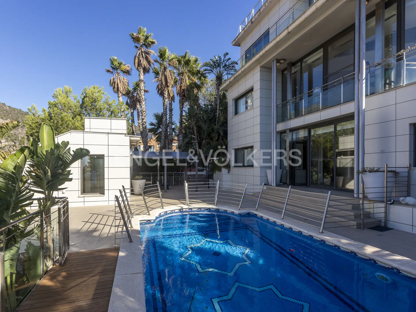 Exterior view of a modern, multi-story white house with a blue mosaic-tiled pool, palm trees, and a clear blue sky.