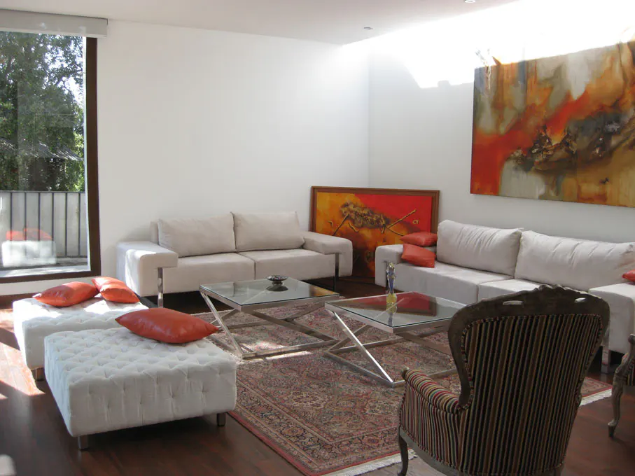 Bright living room with white walls, hardwood floors, and neutral furniture. Two sofas, ottomans with orange pillows, and abstract art.