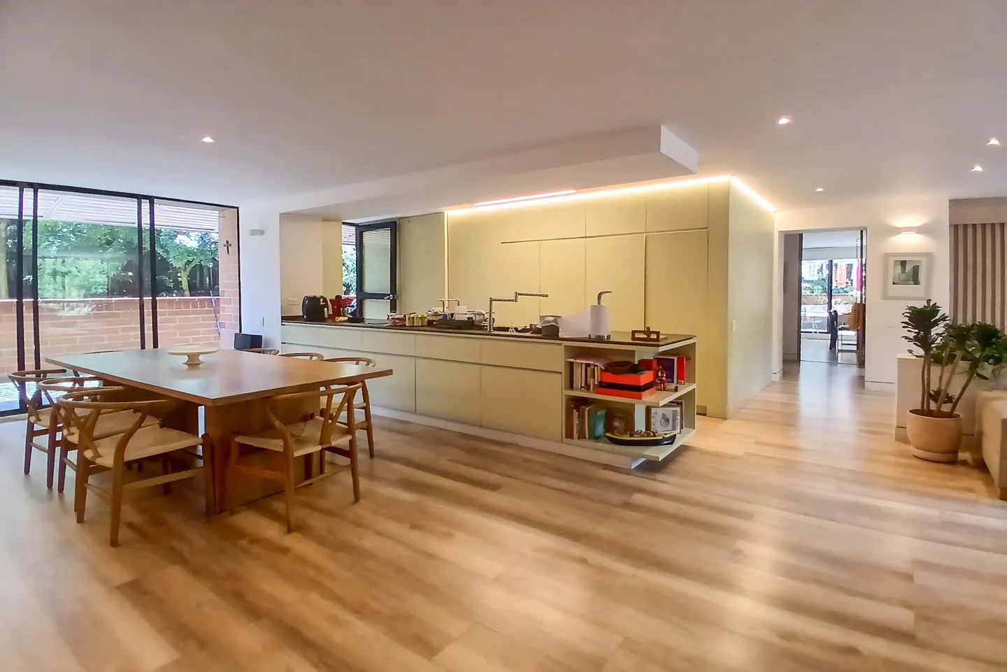 Open-concept kitchen and dining area with light wood floors. A square wooden table with chairs sits near a large window. The kitchen has pale green cabinets.