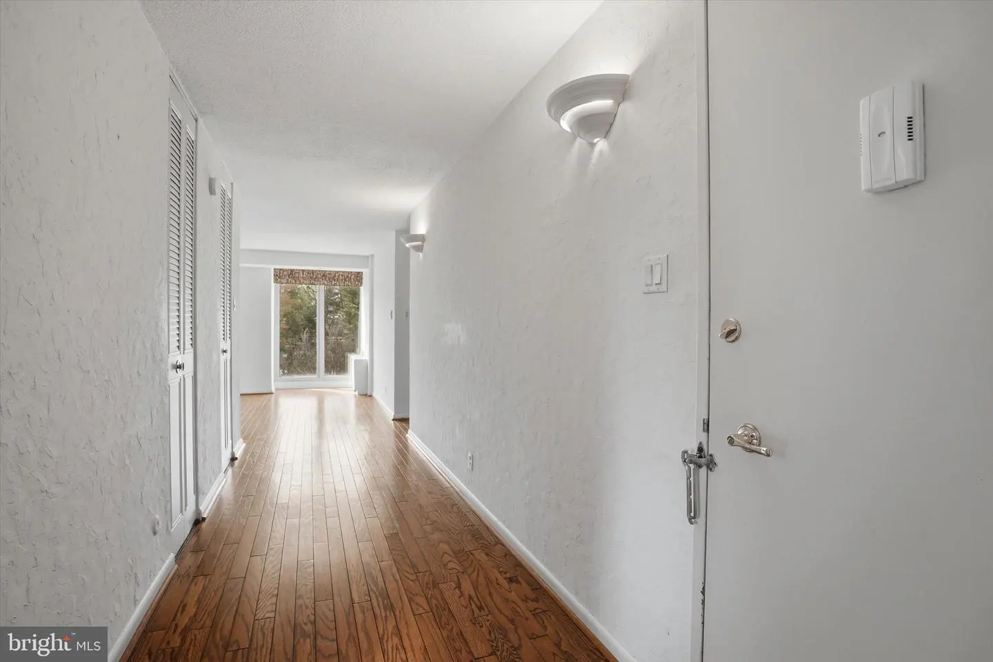 Hallway with wood floors, white walls, and a front door. A window is at the end of the hall.