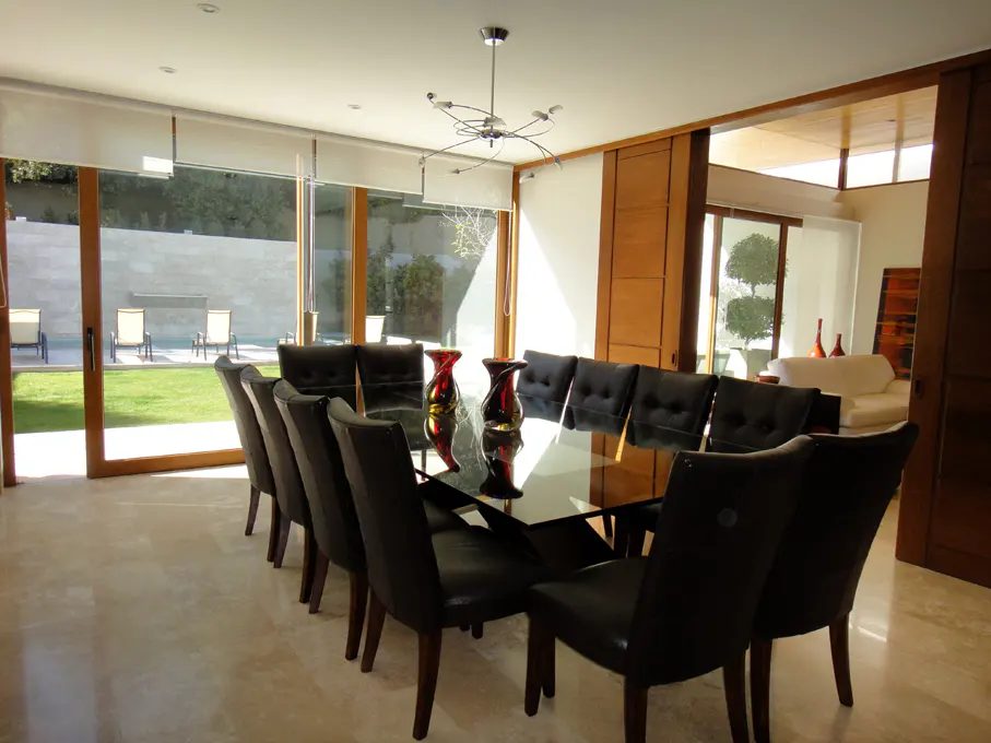 A modern dining room with a glass table, black chairs, and a view of a green lawn with lounge chairs.