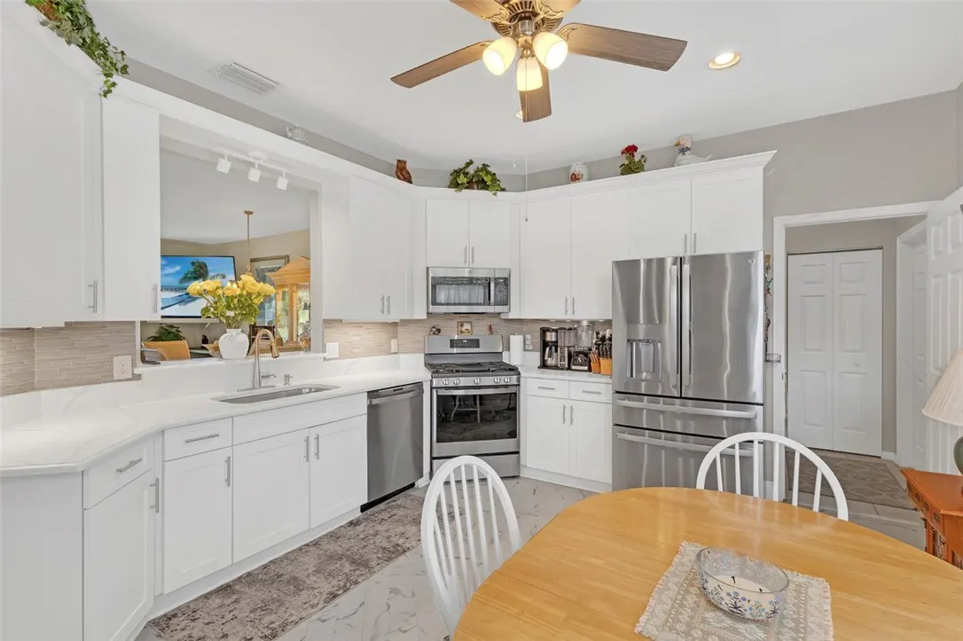 Bright kitchen with white cabinets, stainless steel appliances, and a wood table. A ceiling fan hangs above.