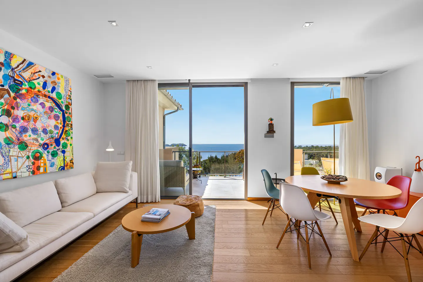 Bright living room with white sofa, colorful art, wood floors, and a view of the ocean through sliding glass doors.