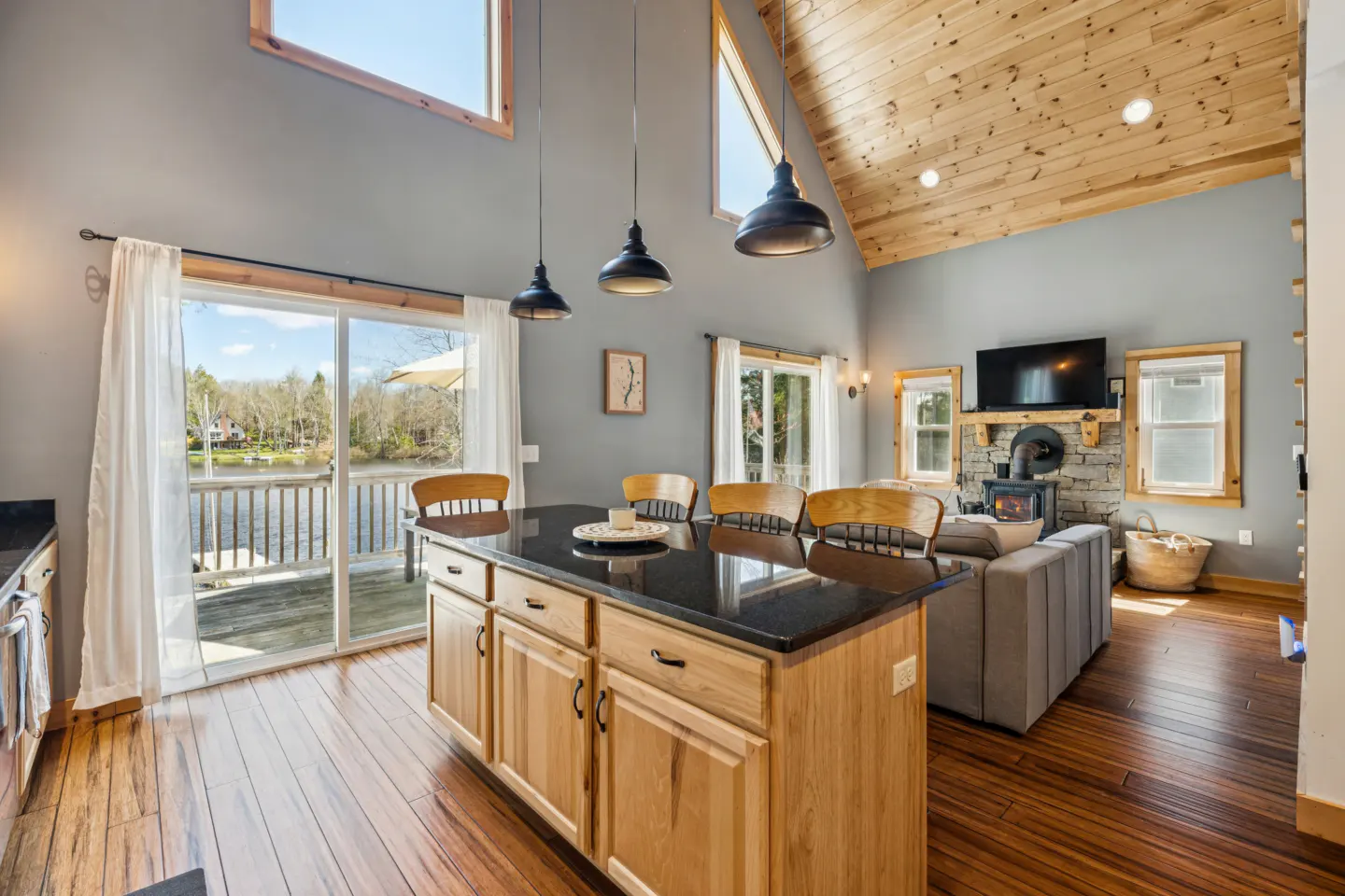 Open-concept kitchen and living room with wood floors, gray walls, and a wood-paneled ceiling. A kitchen island with a black countertop is in the foreground. A lake is visible through sliding glass doors.