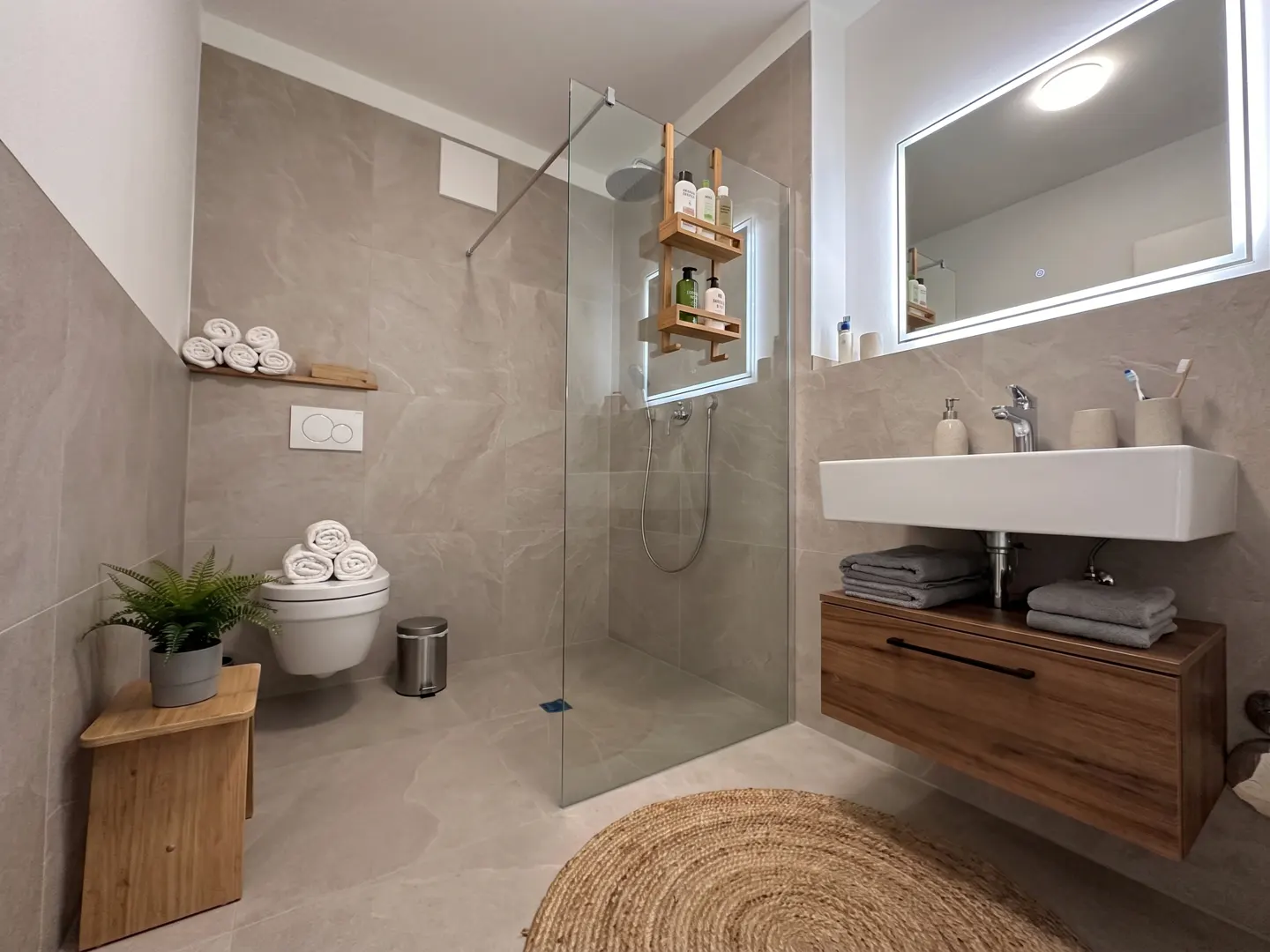 Modern bathroom with gray marble walls, a glass shower, a white sink with a wood cabinet, and a round jute rug.