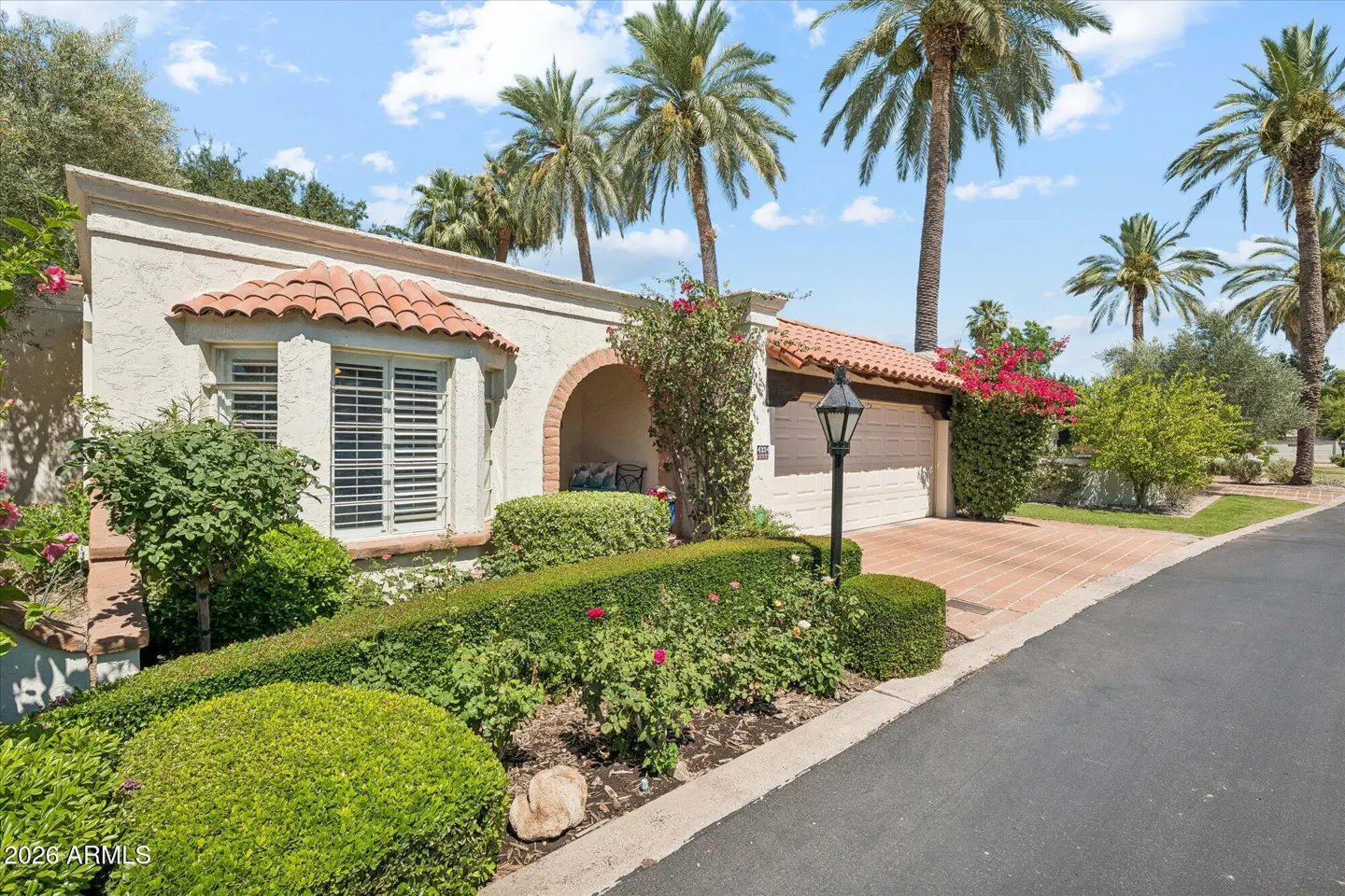 Beige stucco house with red tile roof, arched entry, and garage. Palm trees and manicured green bushes surround the property.