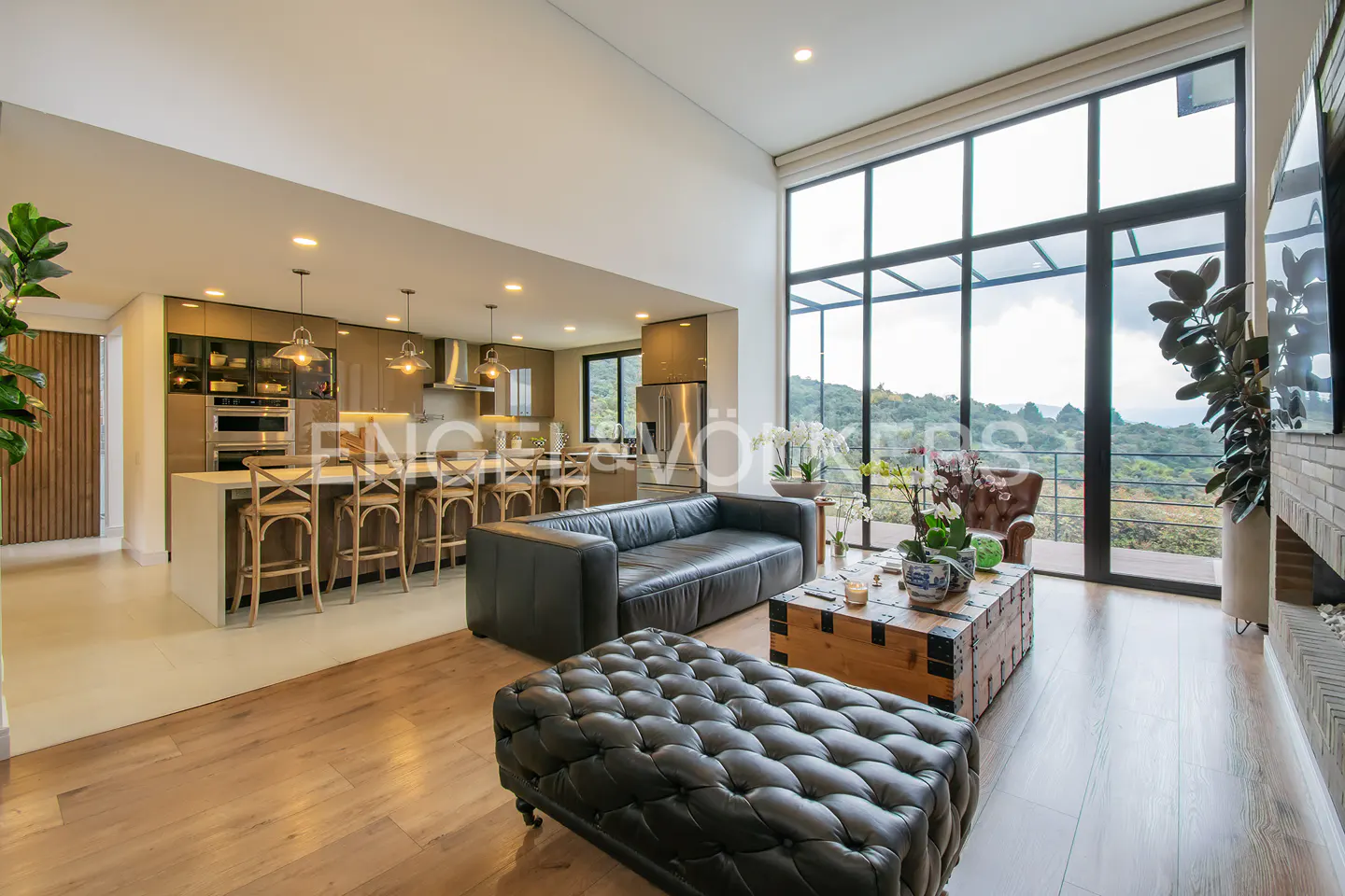 Open-concept living space with a black leather sofa, tufted ottoman, and a kitchen island with wooden stools. Large windows offer a view of lush greenery.