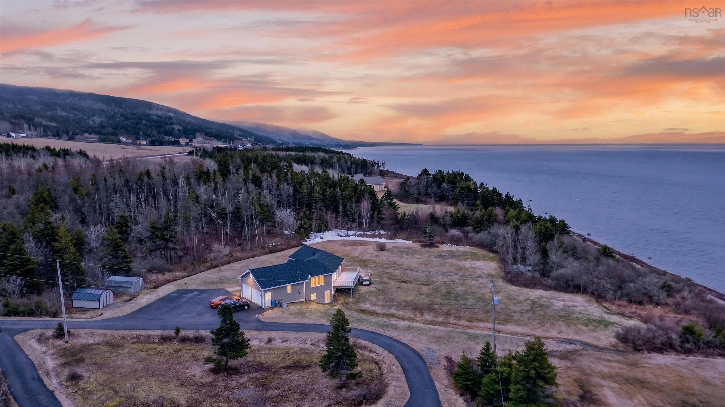 Aerial view of a tan house with a dark roof, cars in the driveway, and the ocean in the background at sunset.