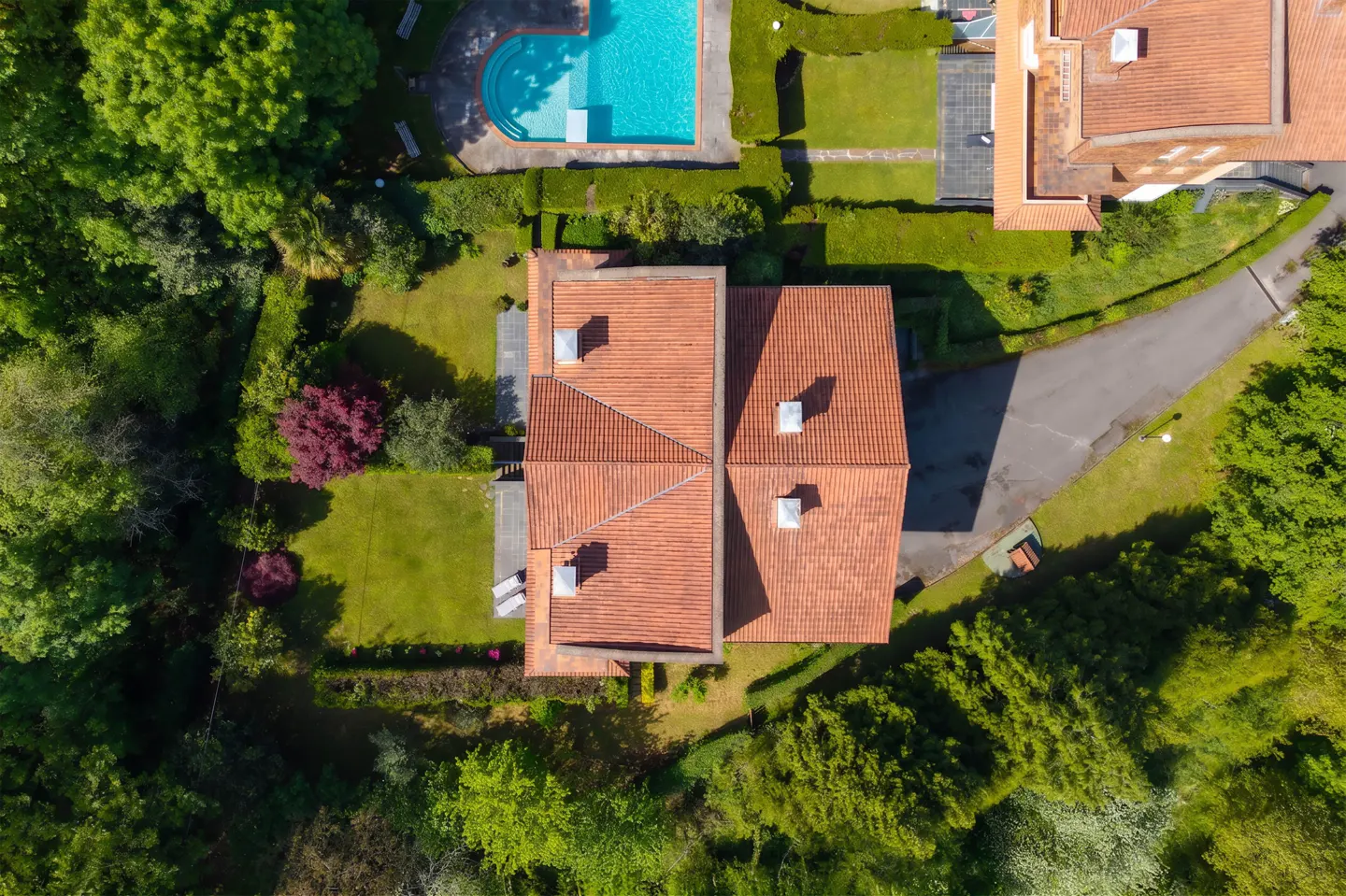 Aerial view of a house with a red tile roof, surrounded by green trees, grass, and a blue swimming pool.