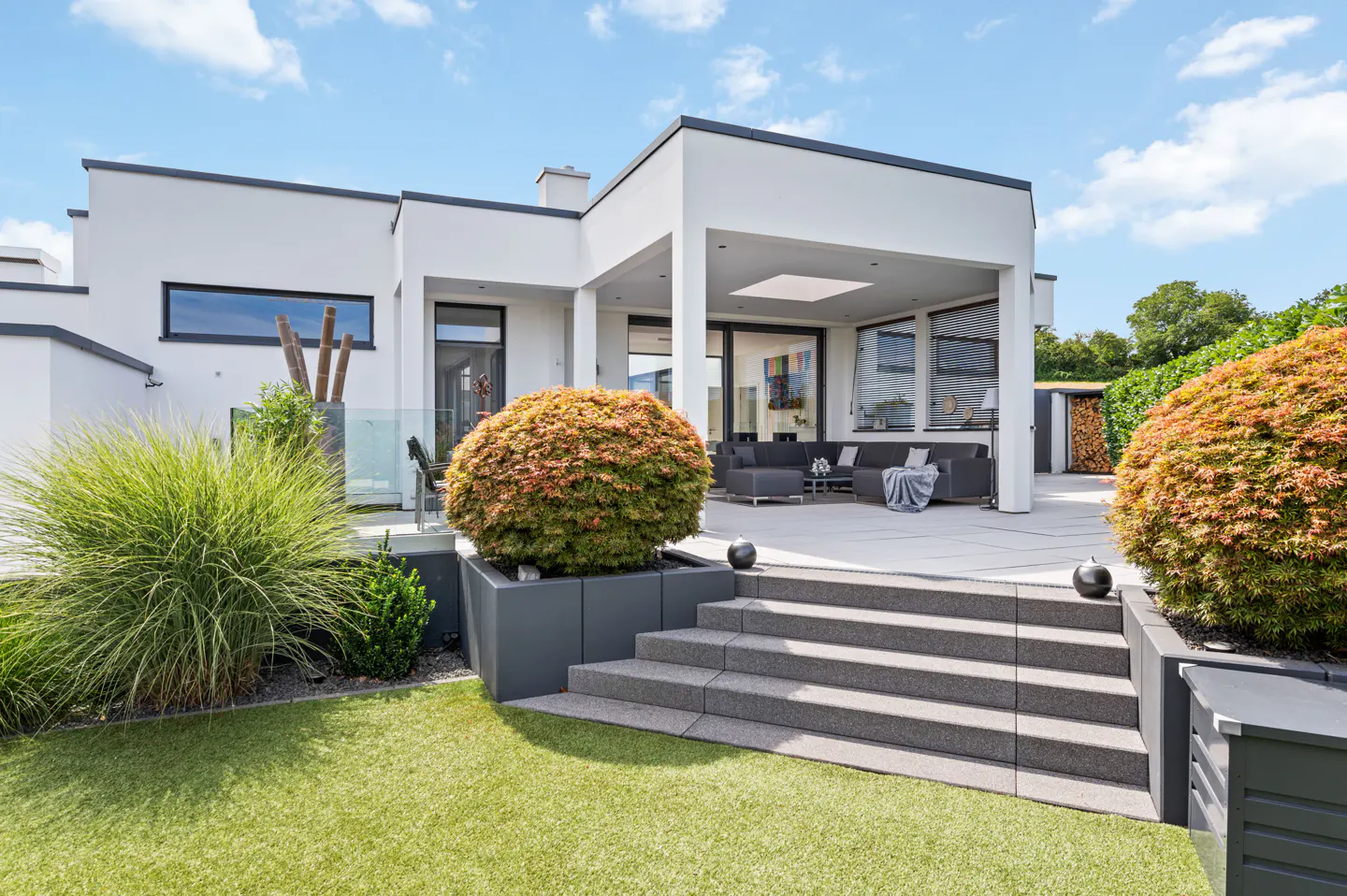 Modern white house with a gray patio, steps, and planters. Green lawn and bushes surround the house. Blue sky with clouds above.