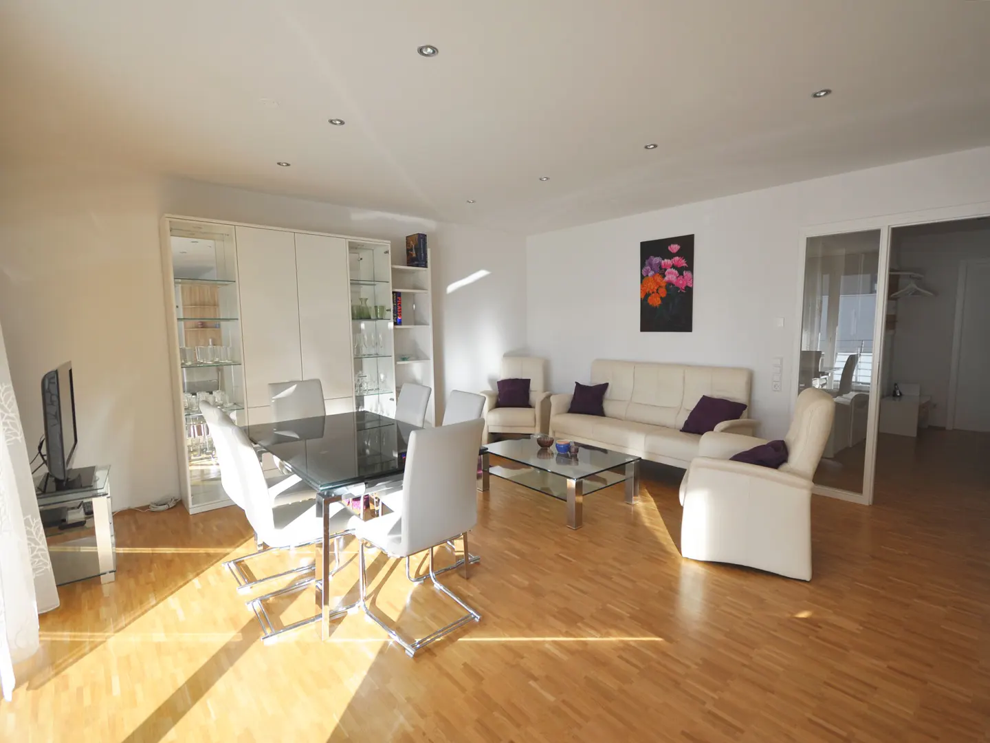 Bright, modern living room with a glass dining table, white leather sofa and chairs, and light wood floors. A colorful floral painting hangs on the wall.