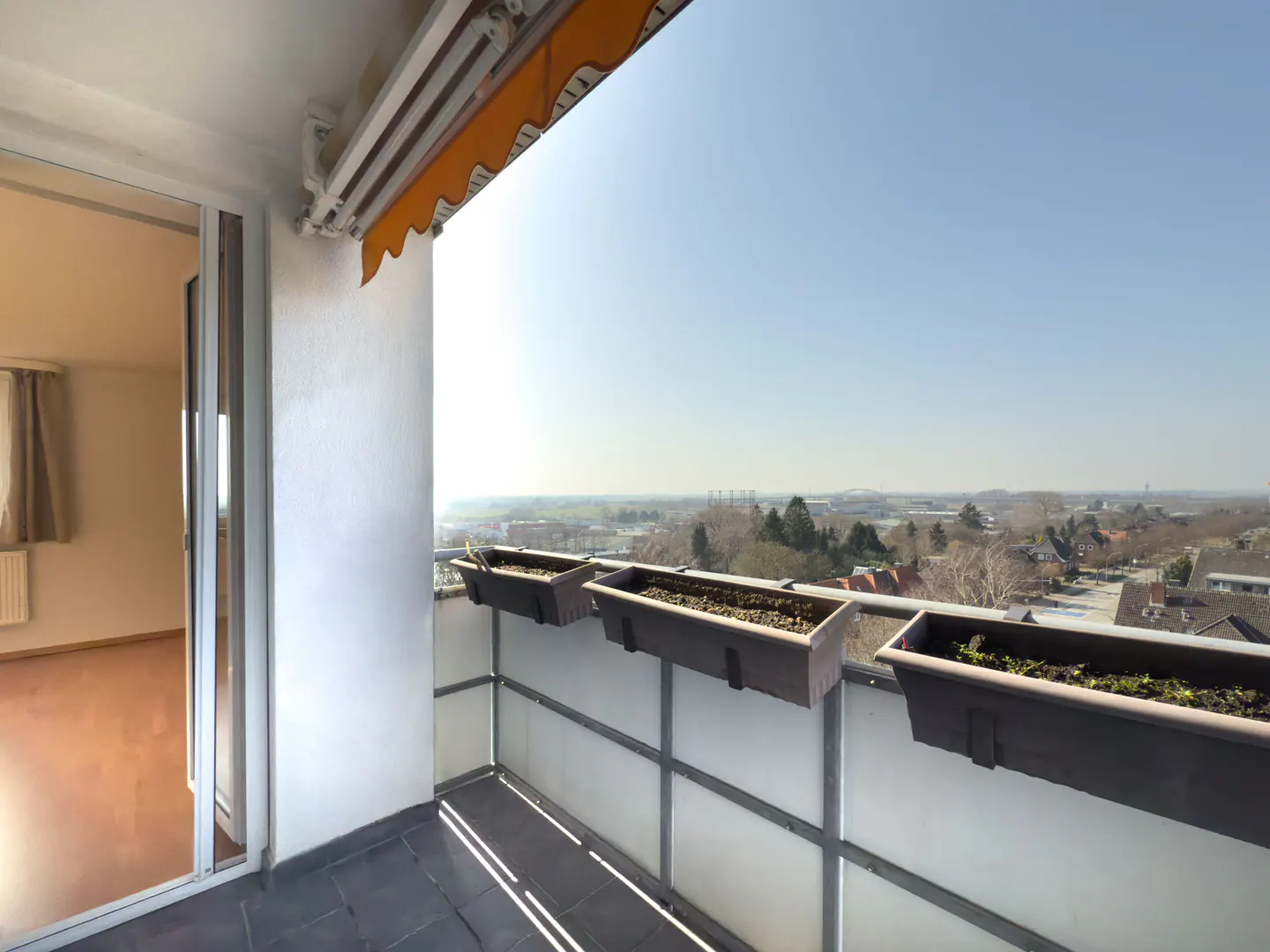 Balcony view with flower boxes, open sliding glass door to room with wood floor. Cityscape in background under a clear blue sky.