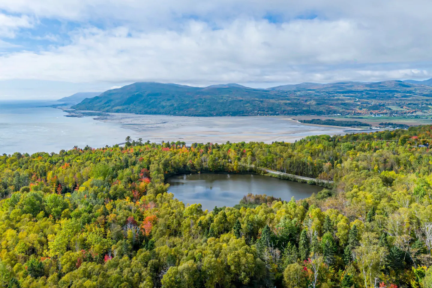 Aerial view of a lake surrounded by colorful fall foliage, with mountains and a body of water in the background.