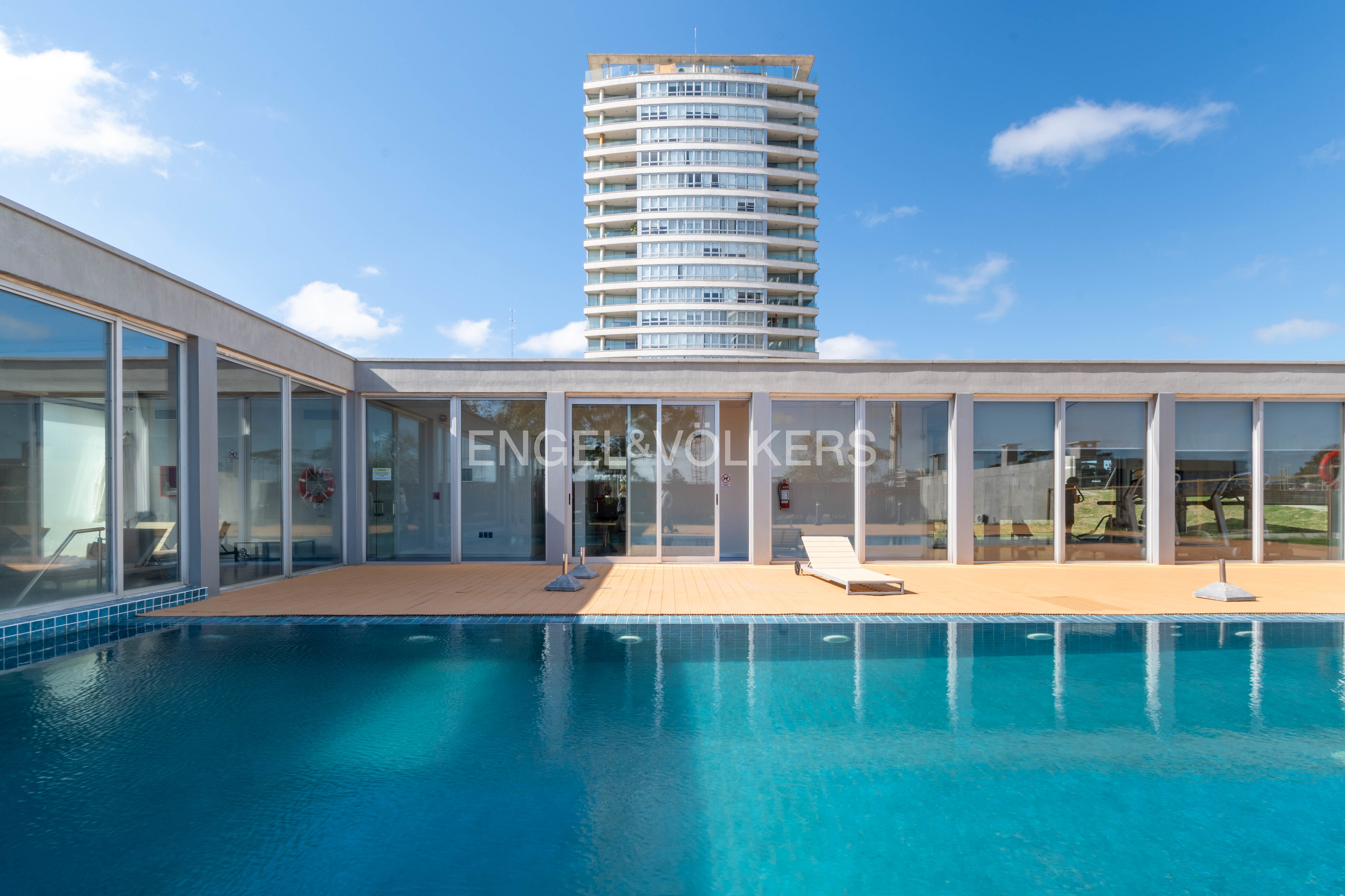 Swimming pool with blue water in front of a building with the Engel & Völkers logo and a tall apartment building in the background.