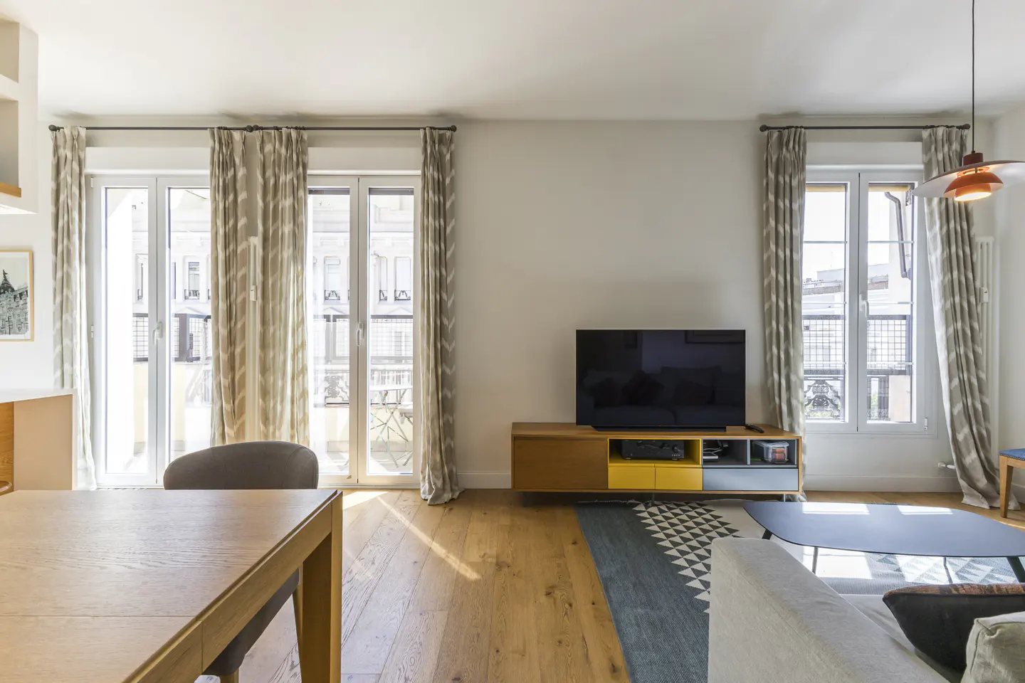 Bright living room with wood floors, large windows with patterned curtains, TV on a yellow and wood stand, and a geometric rug.