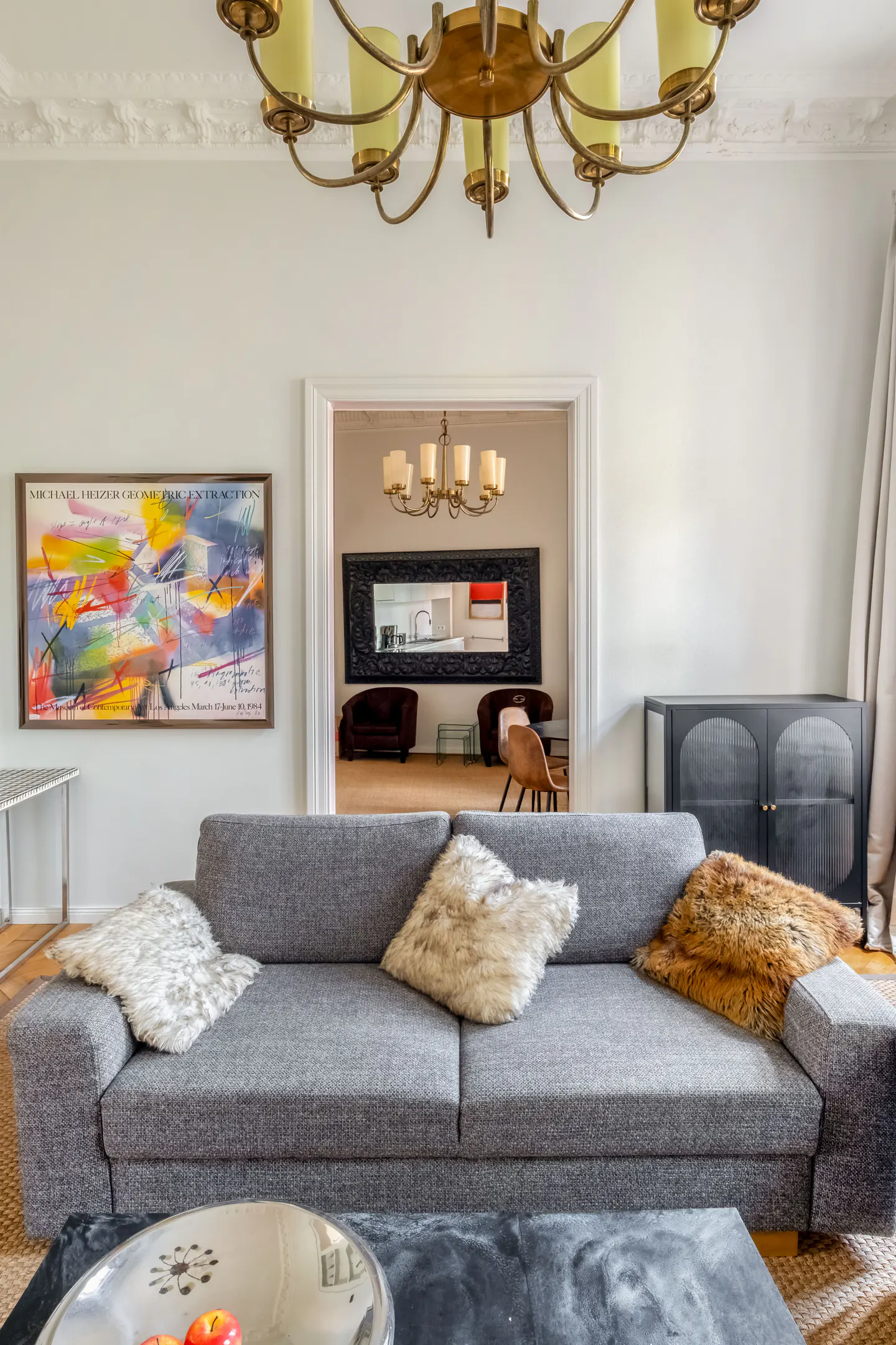 Living room view with a gray sofa, fur pillows, and a colorful abstract painting on the wall. A doorway leads to another room with a mirror and furniture.