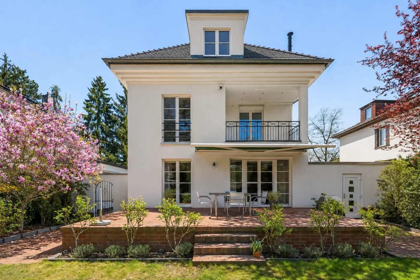 Two-story white house with a balcony, a brick patio with a table and chairs, and a green lawn. Trees and flowers surround the house.