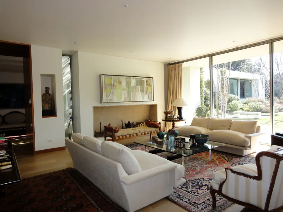 Bright living room with white sofas, a glass coffee table, and a large window overlooking a green garden. A fireplace and artwork add warmth.