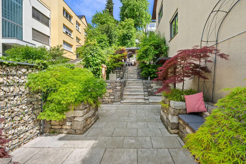 Stone patio with steps leading up to lush greenery and buildings. A bench with a pink pillow sits to the right.