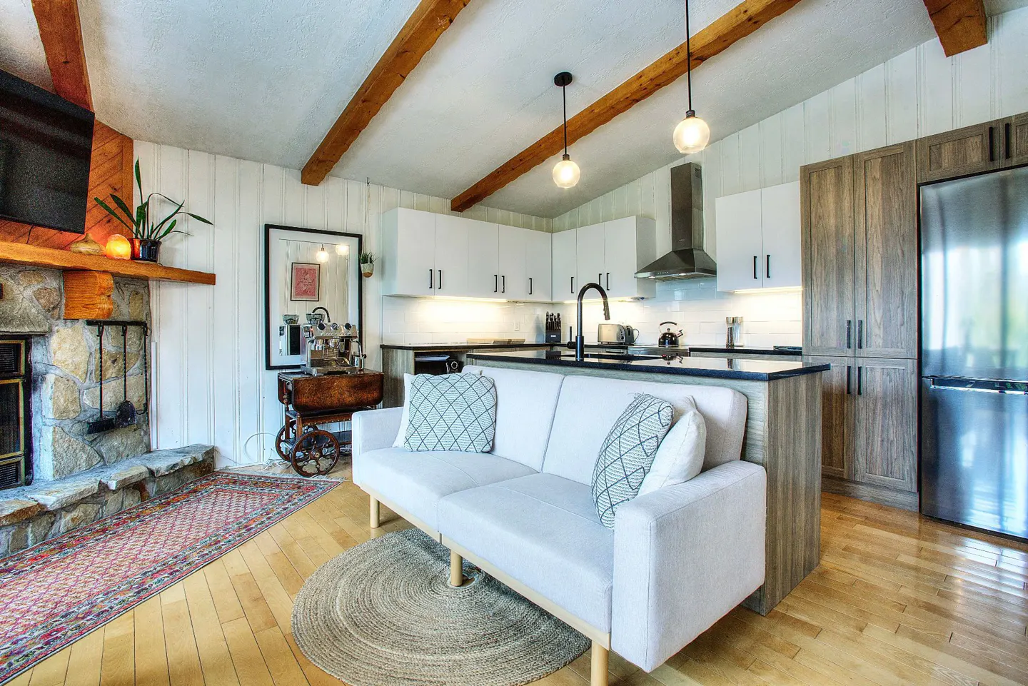 A bright, open-concept living space with a stone fireplace, white sofa, and modern kitchen. Wooden beams accent the ceiling.