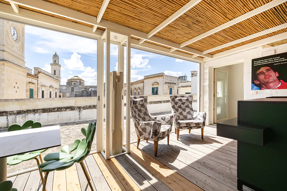 Indoor patio with bamboo ceiling, wooden floor, and open view of a European city. Two patterned armchairs and green chairs are visible.