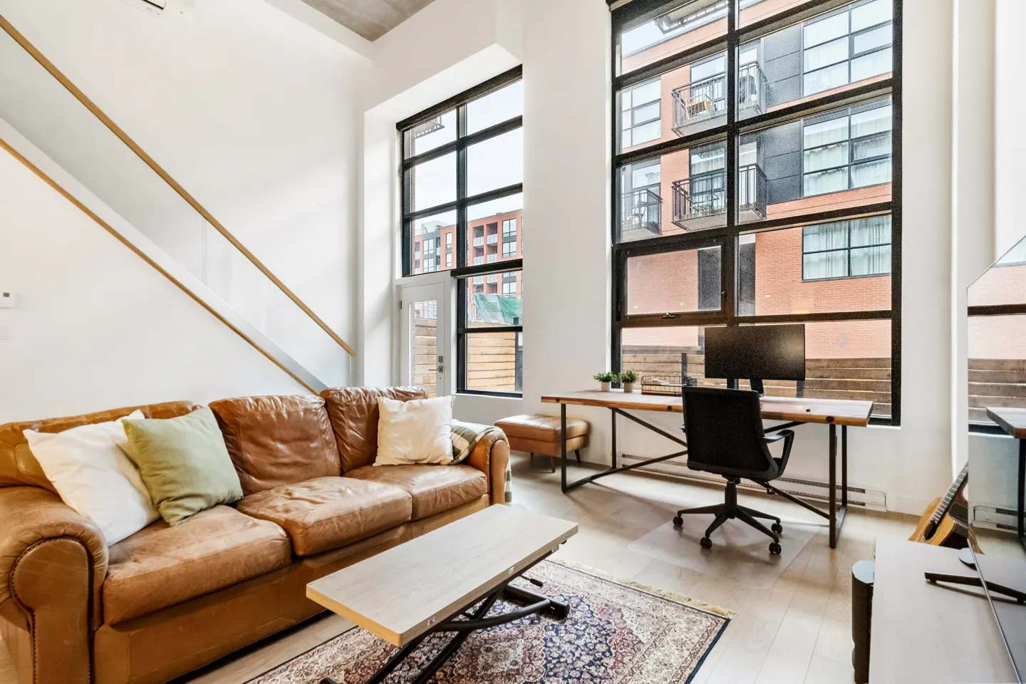 Bright living room with a brown leather sofa, a wooden desk with a computer, and large windows.