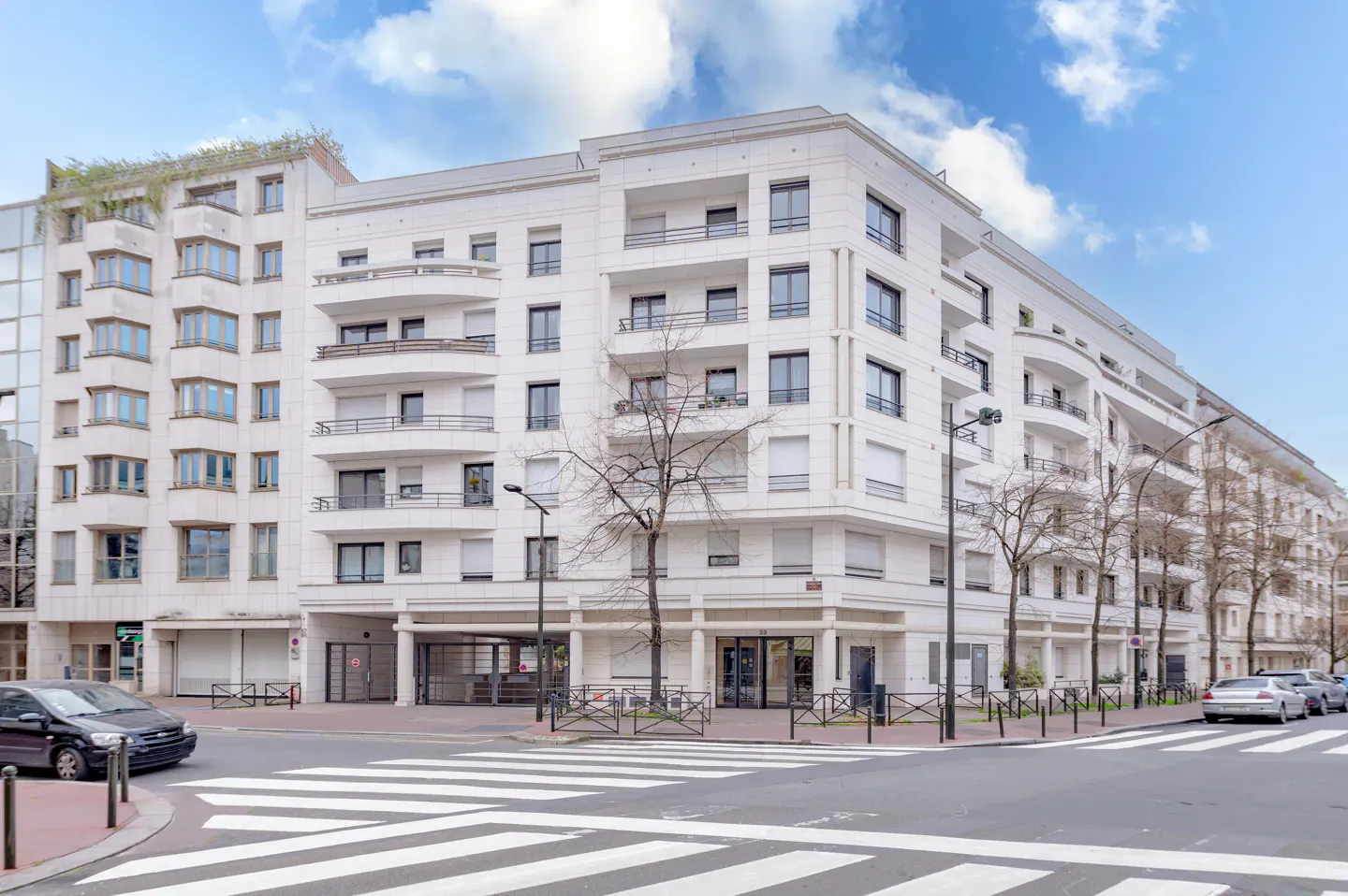 Modern white multi-story apartment building on a city street corner with a crosswalk and parked cars under a blue sky.