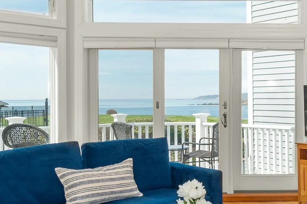 Living room with blue sofa, striped pillow, and white flowers. Patio doors open to a deck with ocean view.