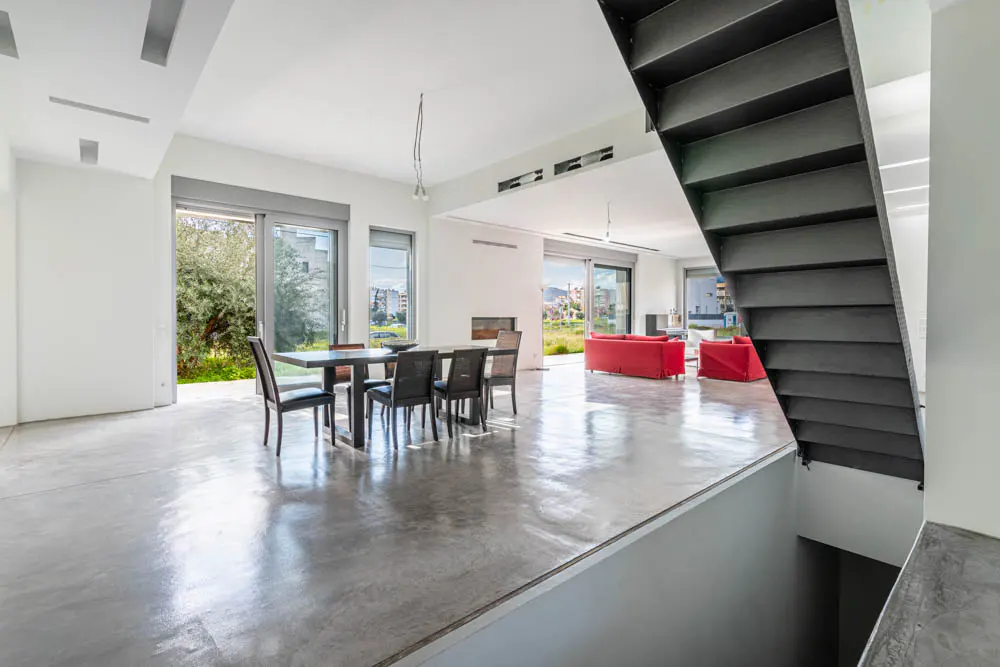 Bright, modern interior with polished concrete floors, dining table, red sofas, and open staircase. Large windows offer outdoor views.